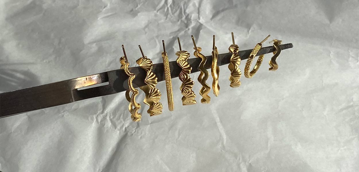 Nine intricately textured gold hoop earrings hanging off a pair of tweezers