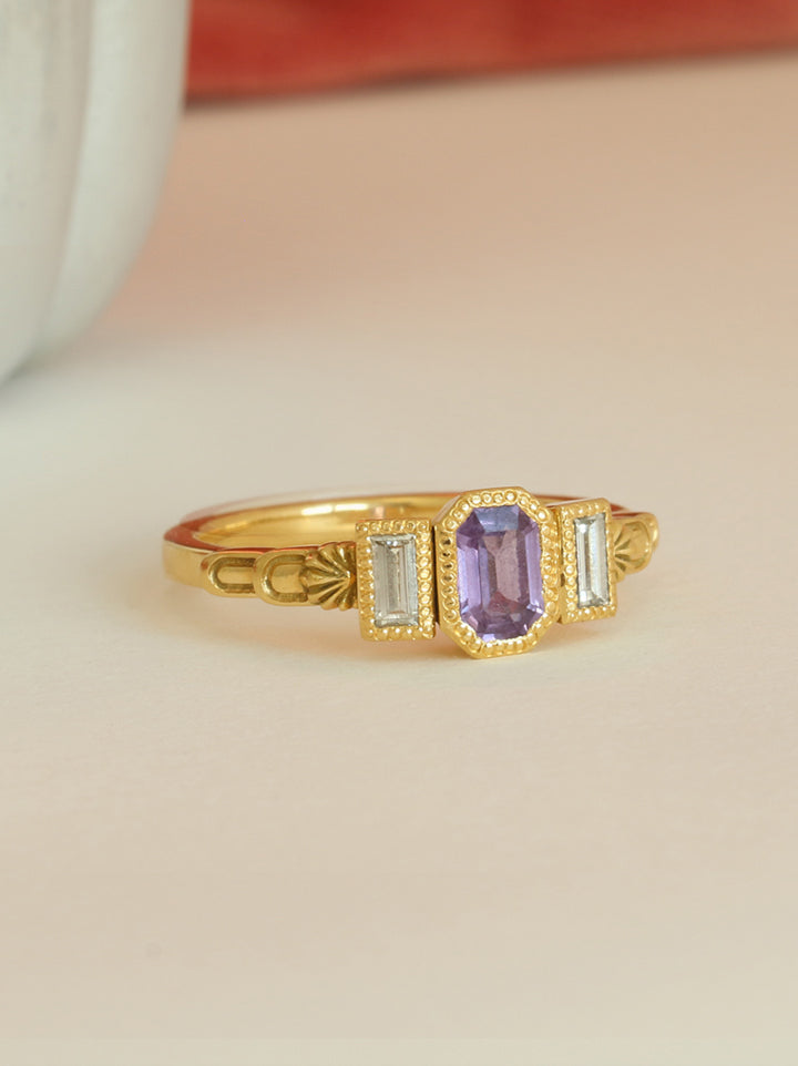 Three stone alternative sea shell engagement ring featuring a central purple emerald cut sapphire and two diamond baguettes either side