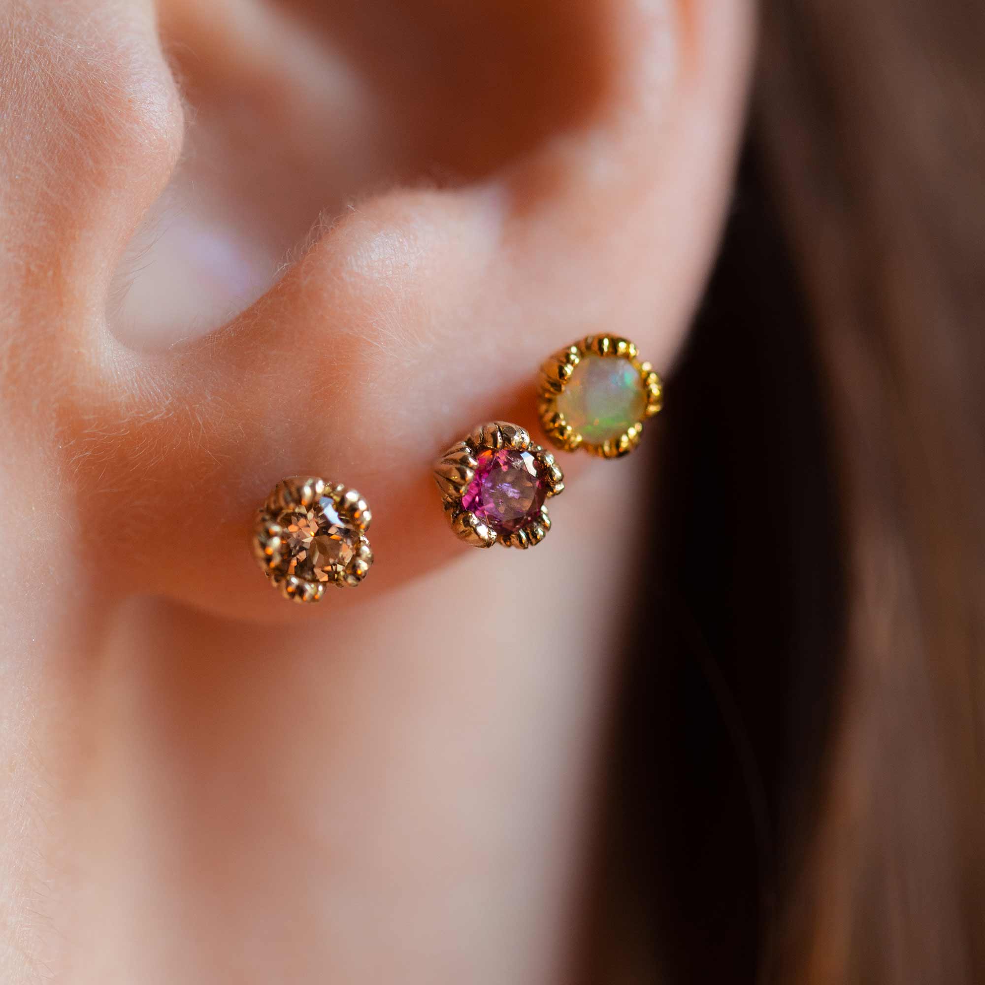Model wears three gemstone stud earrings in one ear. Close up. Showing imperial topaz, pink sapphire and faceted opal.