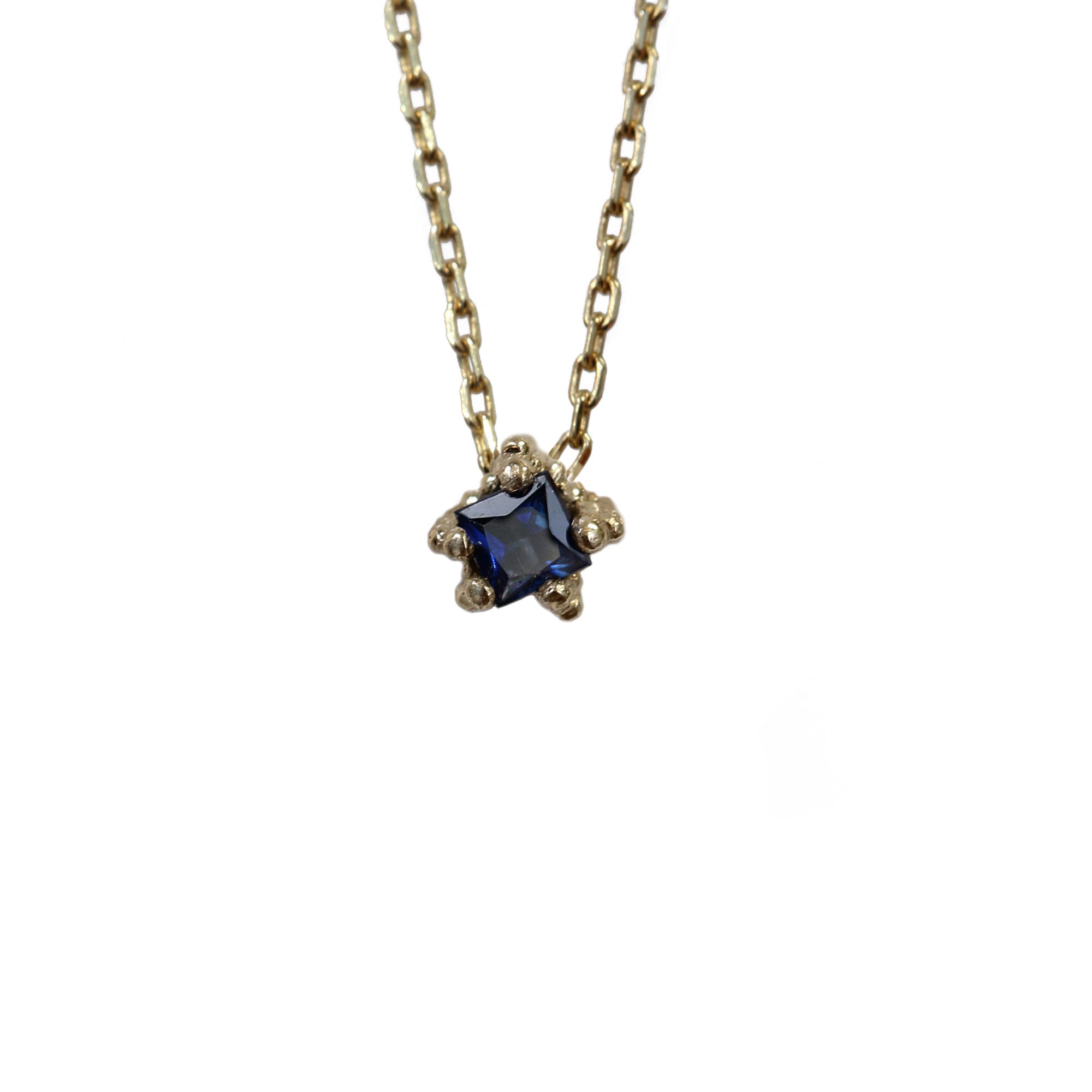 Tiny square cut blue sapphire set in textured gold necklace on white background